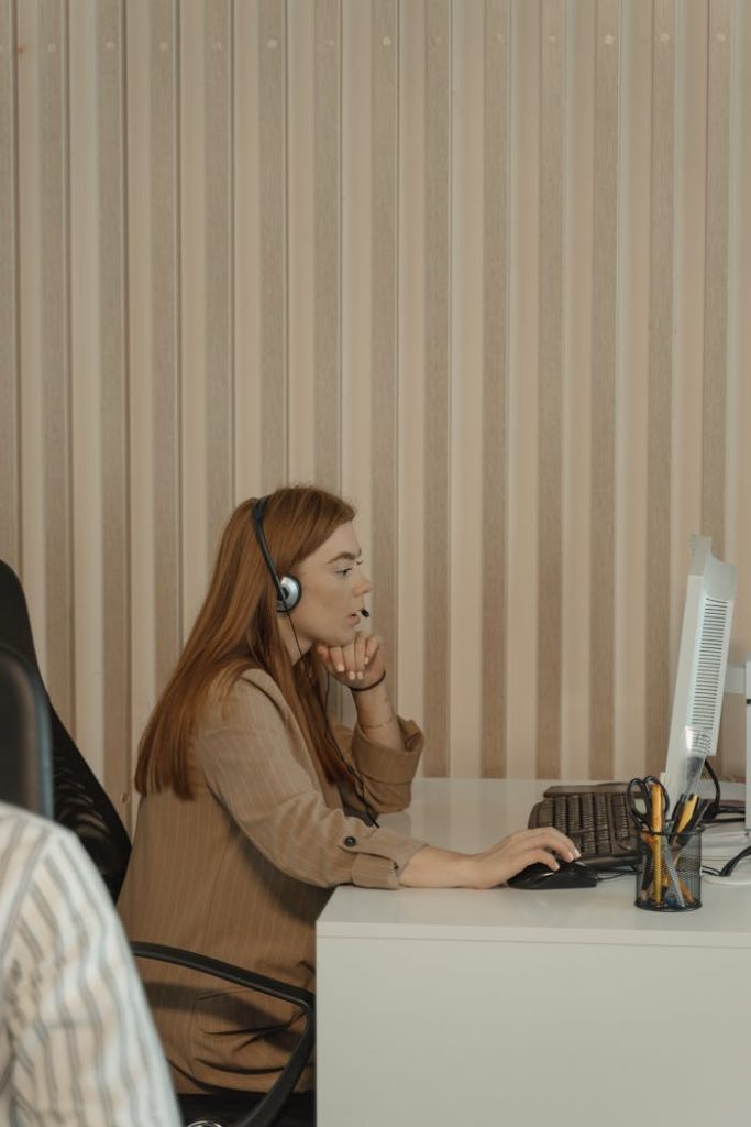 redhead-woman-sitting-by-desk-and-working-5453927 Red-haired woman with headset working at computer desk in office setting.
